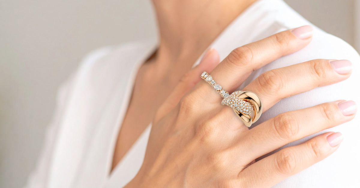 A close-up of a person's hand wearing a gold ring with pavé-set clear stones, paired with a white blouse against a soft neutral background.