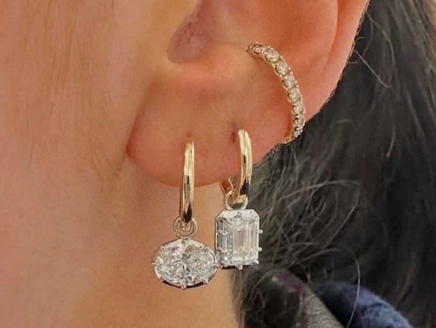 Close-up of a person's ear wearing two gold hoop earrings with clear gemstone drops, one oval and one rectangular emerald-cut style stone, plus a small gold ear cuff.