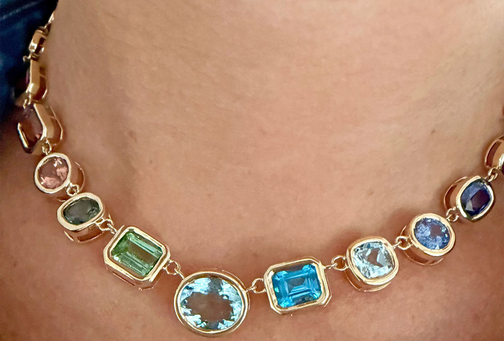 A close-up of a gold necklace worn on a person's neck, featuring multiple bezel-set gemstones in pink, green, light blue, and dark blue.