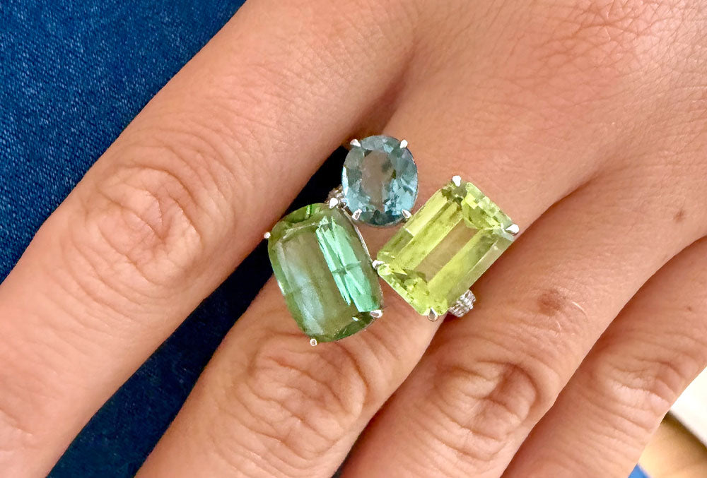 A close-up of a hand wearing a silver ring set with three gemstones: a large emerald-cut green stone, a rectangular yellow-green stone, and a round light blue stone.