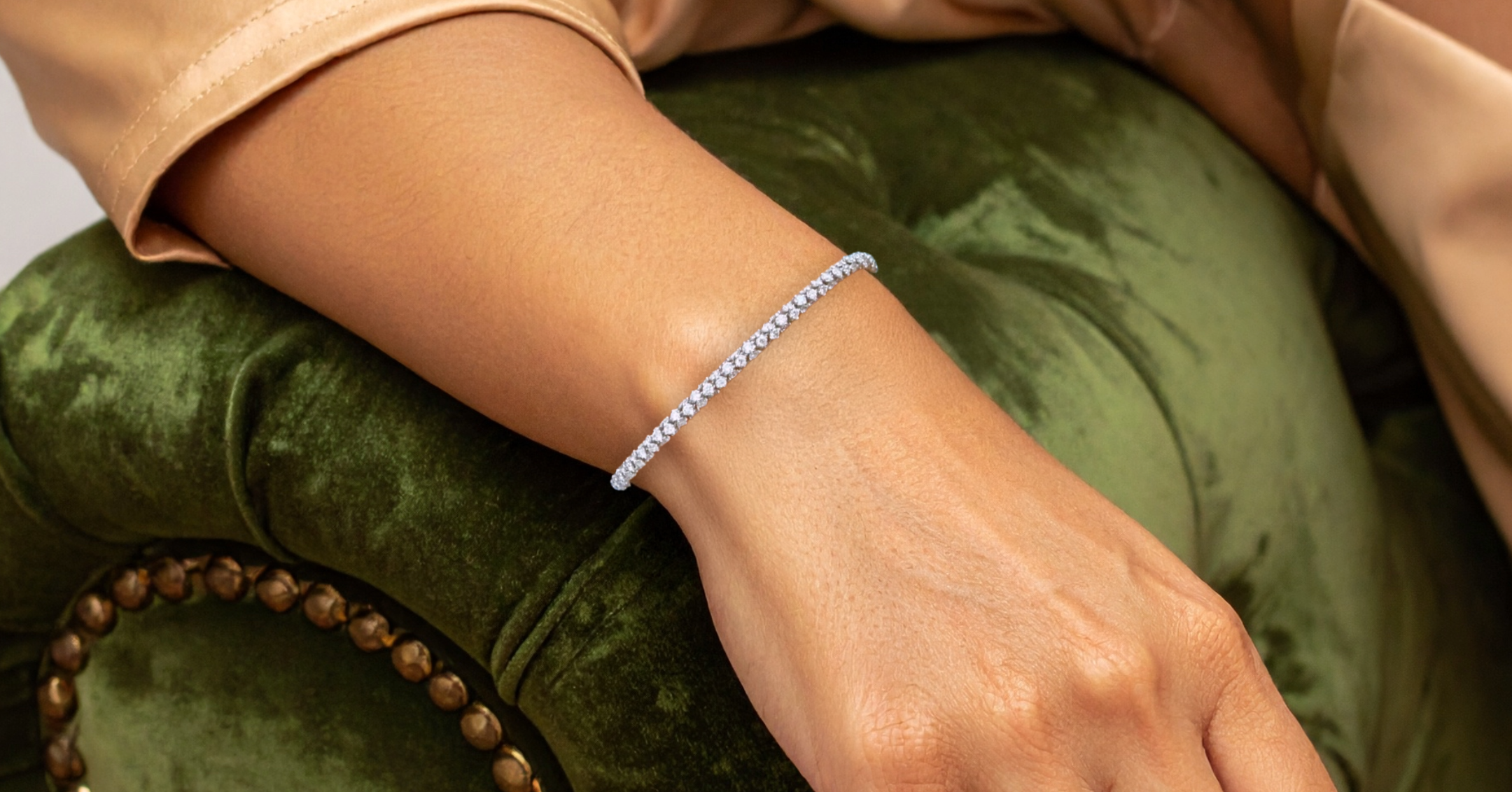 A close-up of a delicate diamond tennis bracelet worn on a woman's wrist, resting on a plush green velvet armchair.