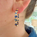 Close-up of a woman wearing 14K yellow gold hoop earrings featuring an organic mosaic of natural blue sapphires accented with round brilliant diamonds.