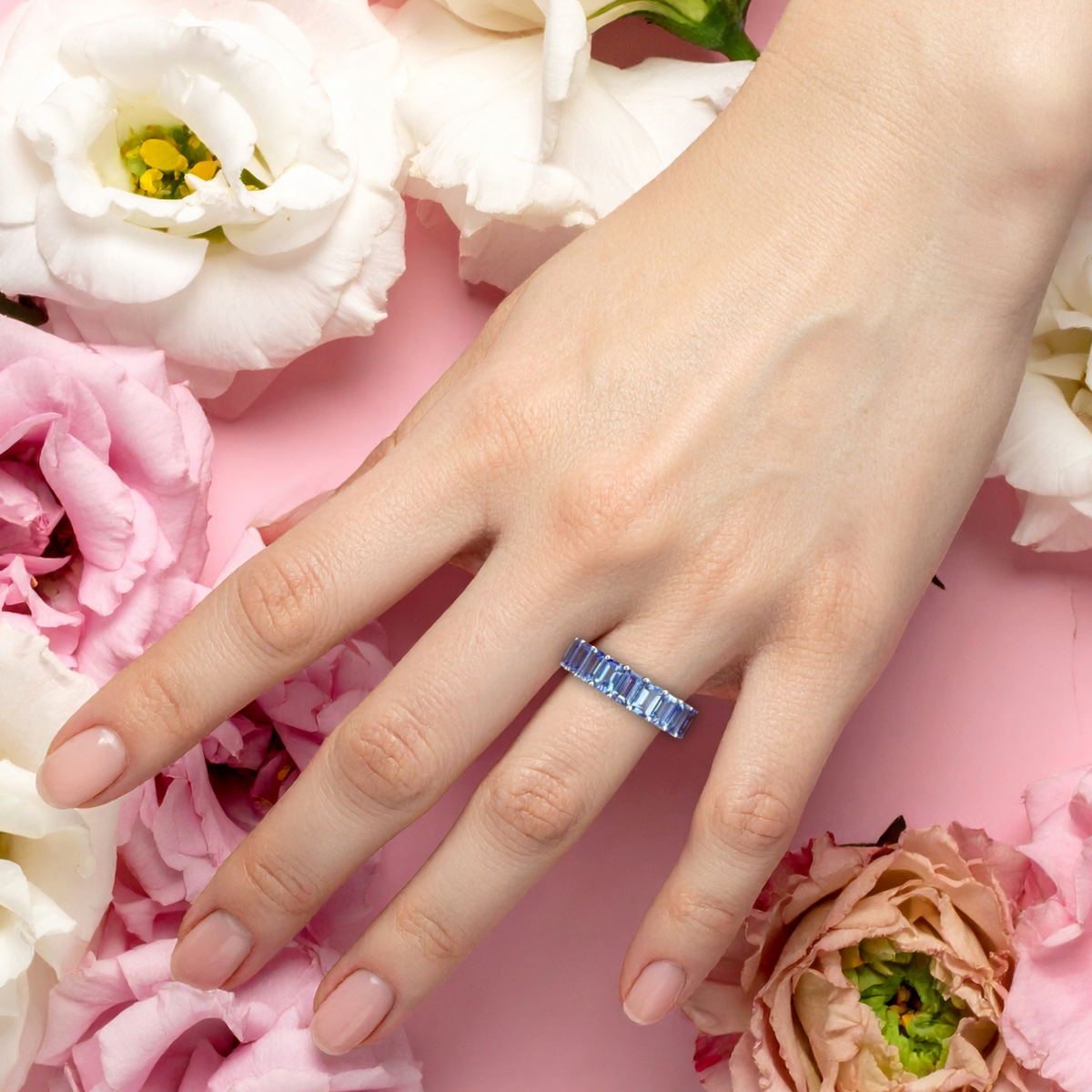 A close-up of a woman's hand wearing a silver band ring set with emerald-cut tanzanite, styled with pink and white flowers on a pink background.