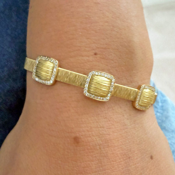 A gold bangle bracelet with three textured square links, each bordered by pave diamonds, shown worn on a wrist.