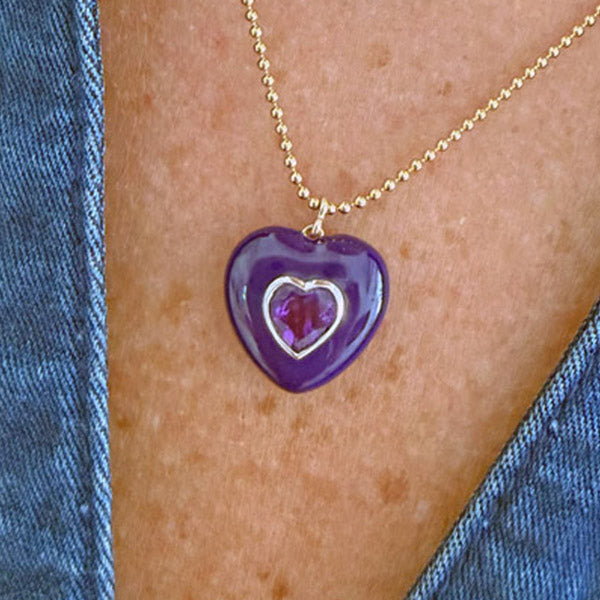 A close-up of a person wearing an amethyst heart-shaped pendant necklace with a smaller inner amethyst heart on a gold ball chain.