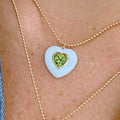 A close-up of a person wearing a white heart-shaped pendant necklace with a smaller inner peridot heart on a gold ball chain.
