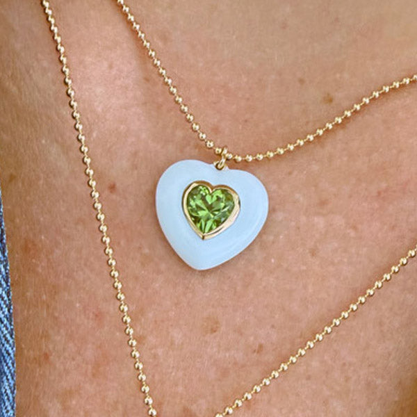 A close-up of a person wearing a white heart-shaped pendant necklace with a smaller inner peridot heart on a gold ball chain.
