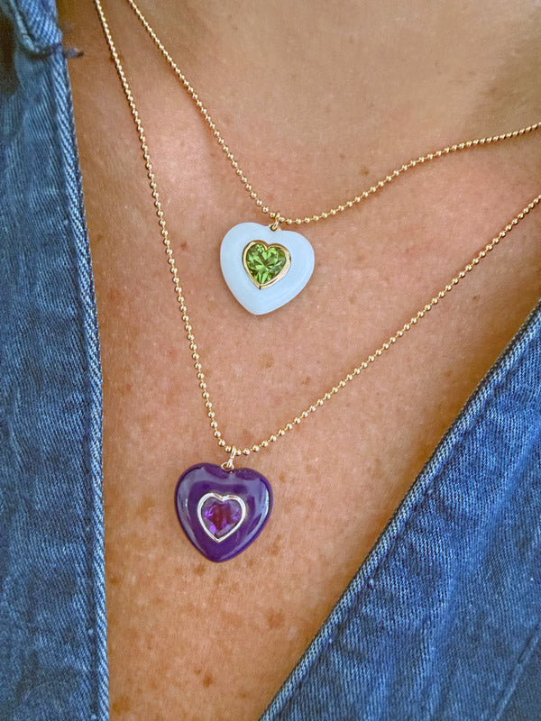 Two layered gold necklaces worn on a model's chest. The top necklace features a white heart-shaped pendant with a green heart inlay.