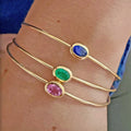 Three slim gold bangle bracelets with oval-cut colored stones (emerald, ruby, and sapphire) worn stacked on a person's wrist.